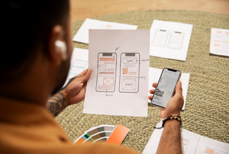 Personne travaillant sur la conception d'une application mobile, tenant un croquis de wireframe dans une main et un smartphone affichant l'application dans l'autre.