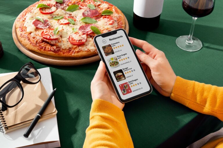 Personne tenant un smartphone avec une application de restaurants ouverte, une pizza appétissante en arrière-plan sur une table.